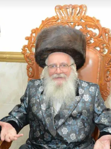 מוהרא״ש יושב על כיסא מגולף בחליפת משי כחולה, כפות ידיו פרושות
