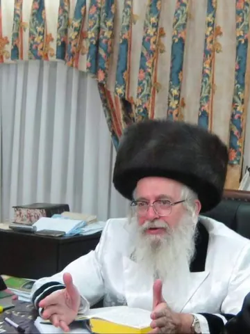 מוהרא״ש בבגדים לבנים מדבר ליד שולחנו, סידור פתוח לפניו